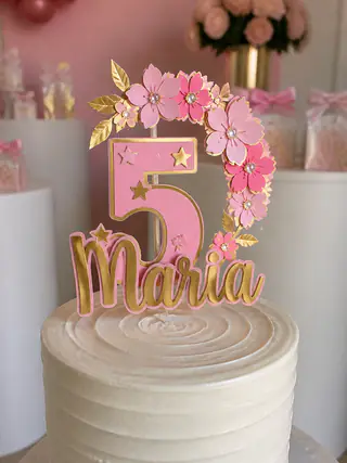 Topo de bolo de papel em rosa e dourado com o nome da aniversariante e flores, espetado em um bolo branco